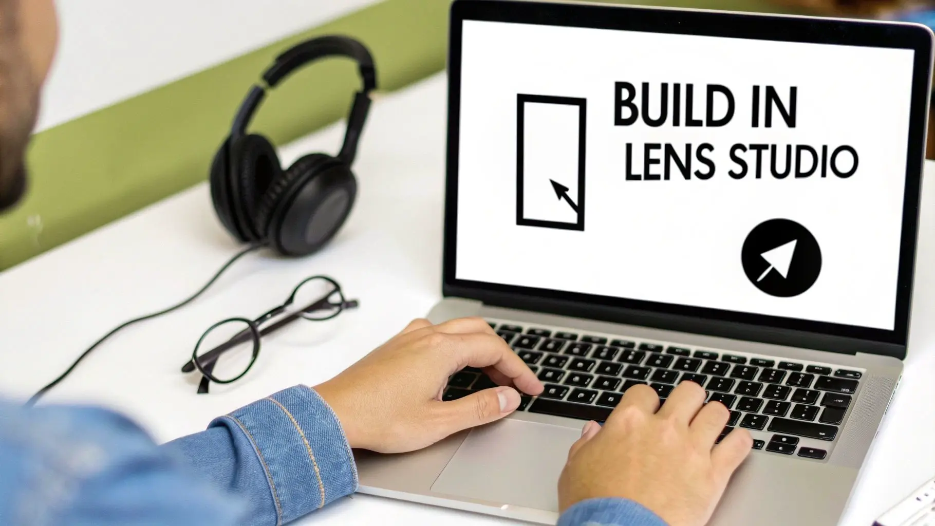 A person types on a laptop displaying BUILD IN LENS STUDIO, with headphones and glasses on the desk.